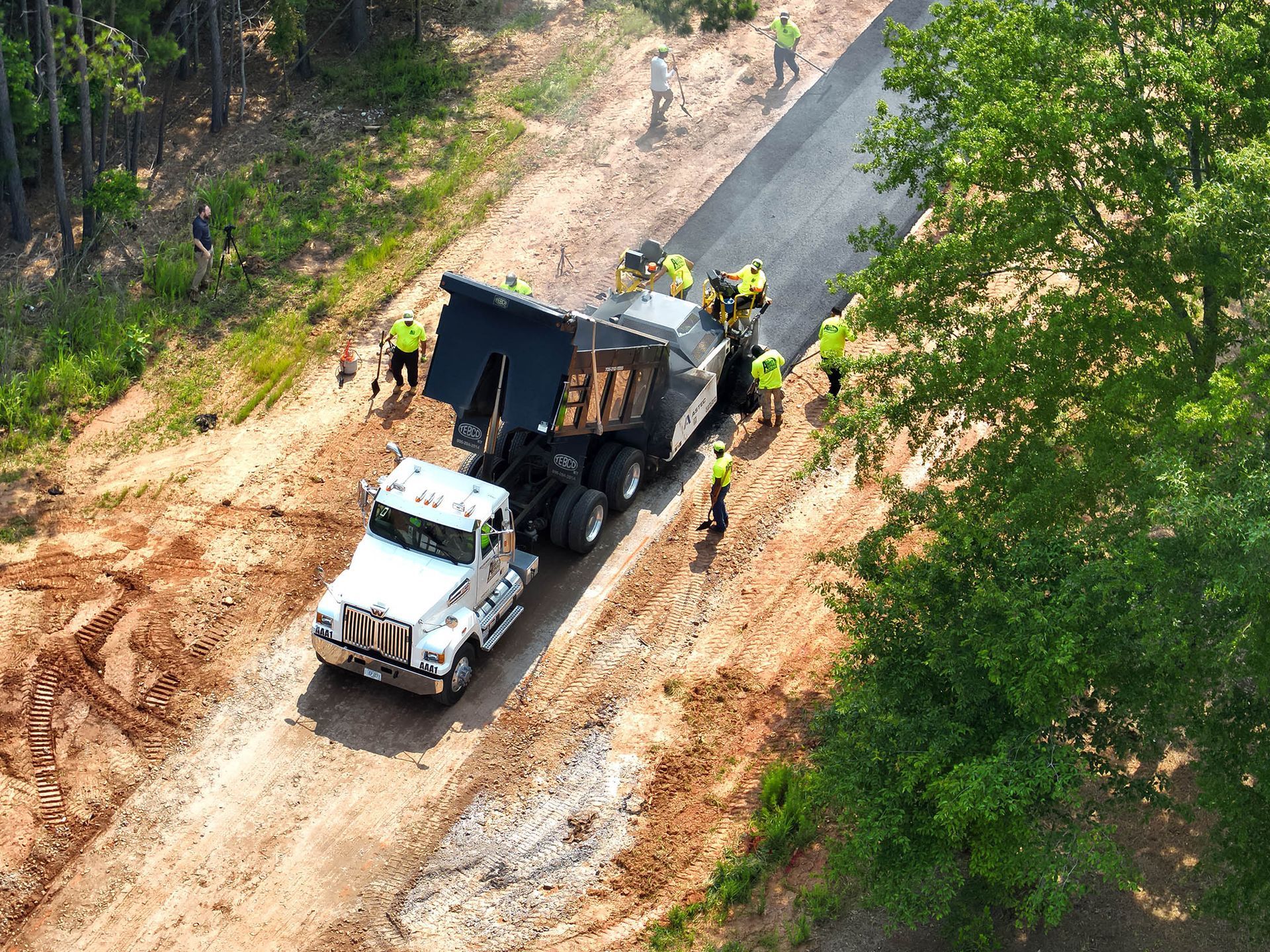 Dump truck unloading asphalt onto a freshly paved road, workers in yellow vests nearby.