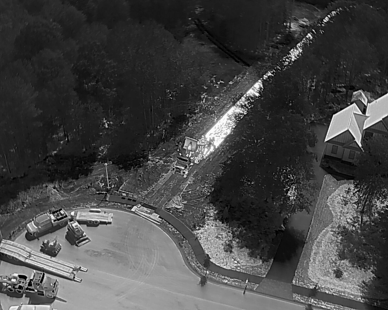 Aerial view of a clearing with a long, bright streak, possibly a river or path, flanked by dark trees and a building.