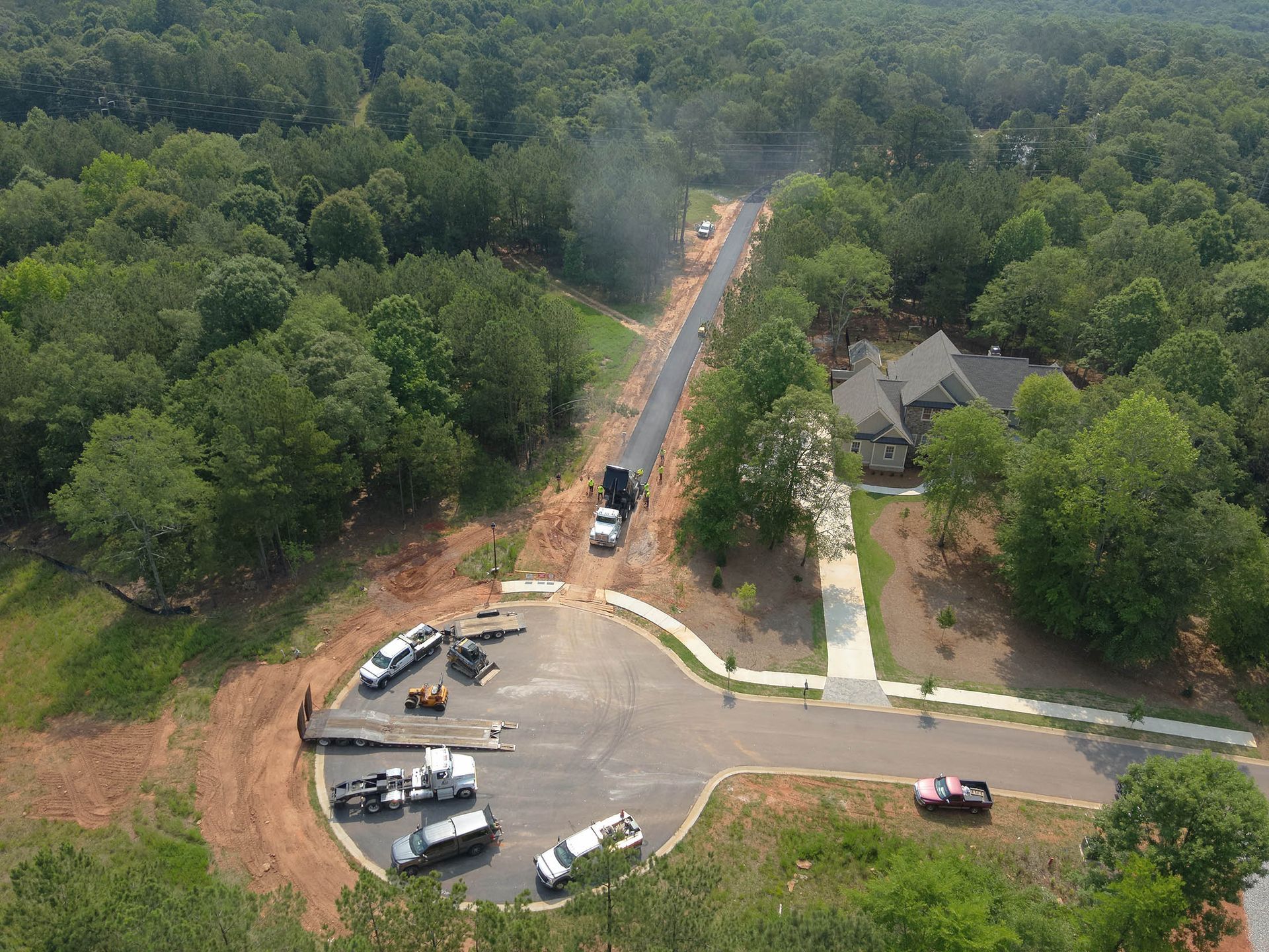 Road construction, aerial view. Asphalt paving with trucks, surrounded by trees and a house.