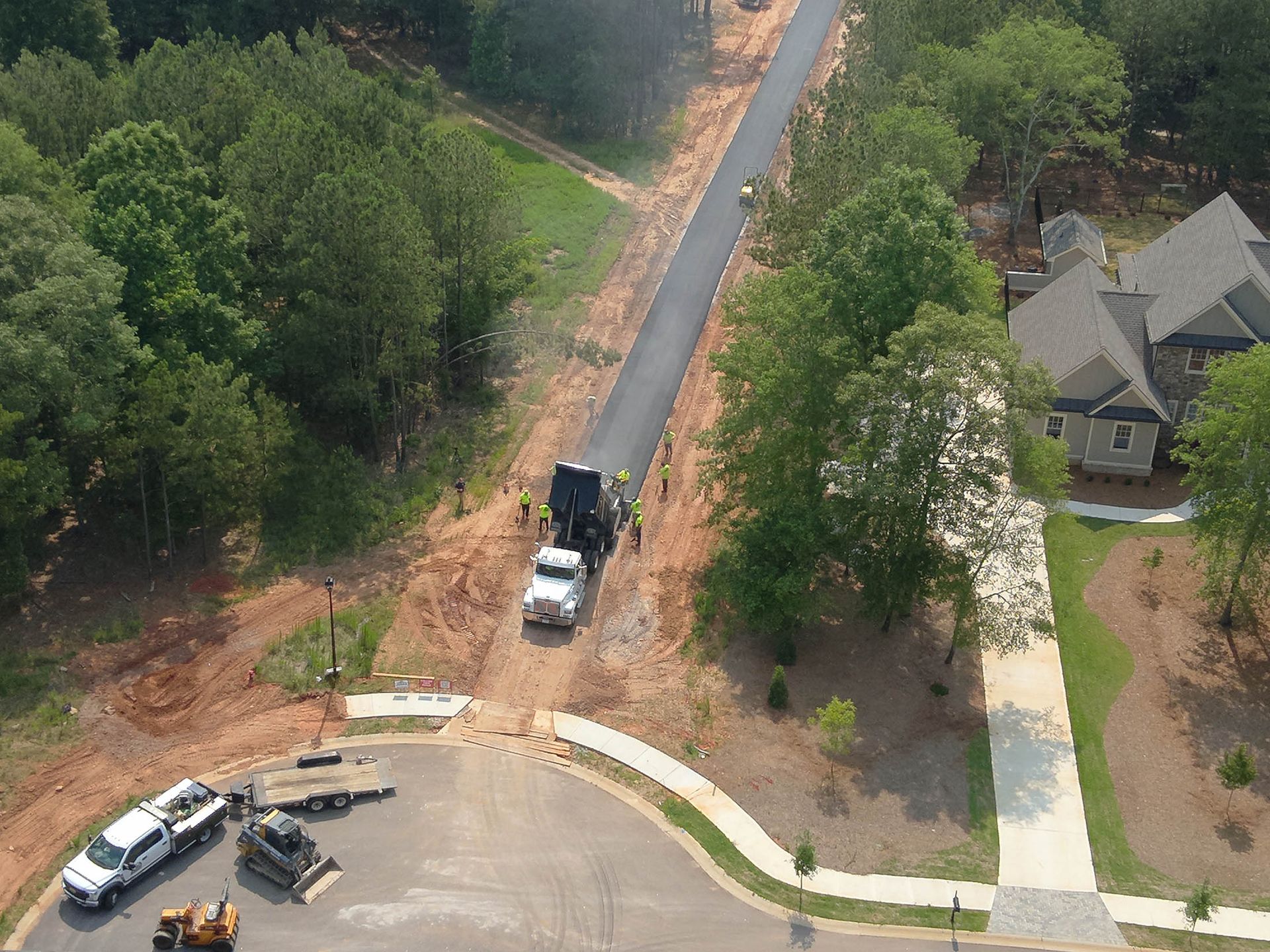 Truck dumping asphalt on new road in a residential area, with equipment and houses visible.