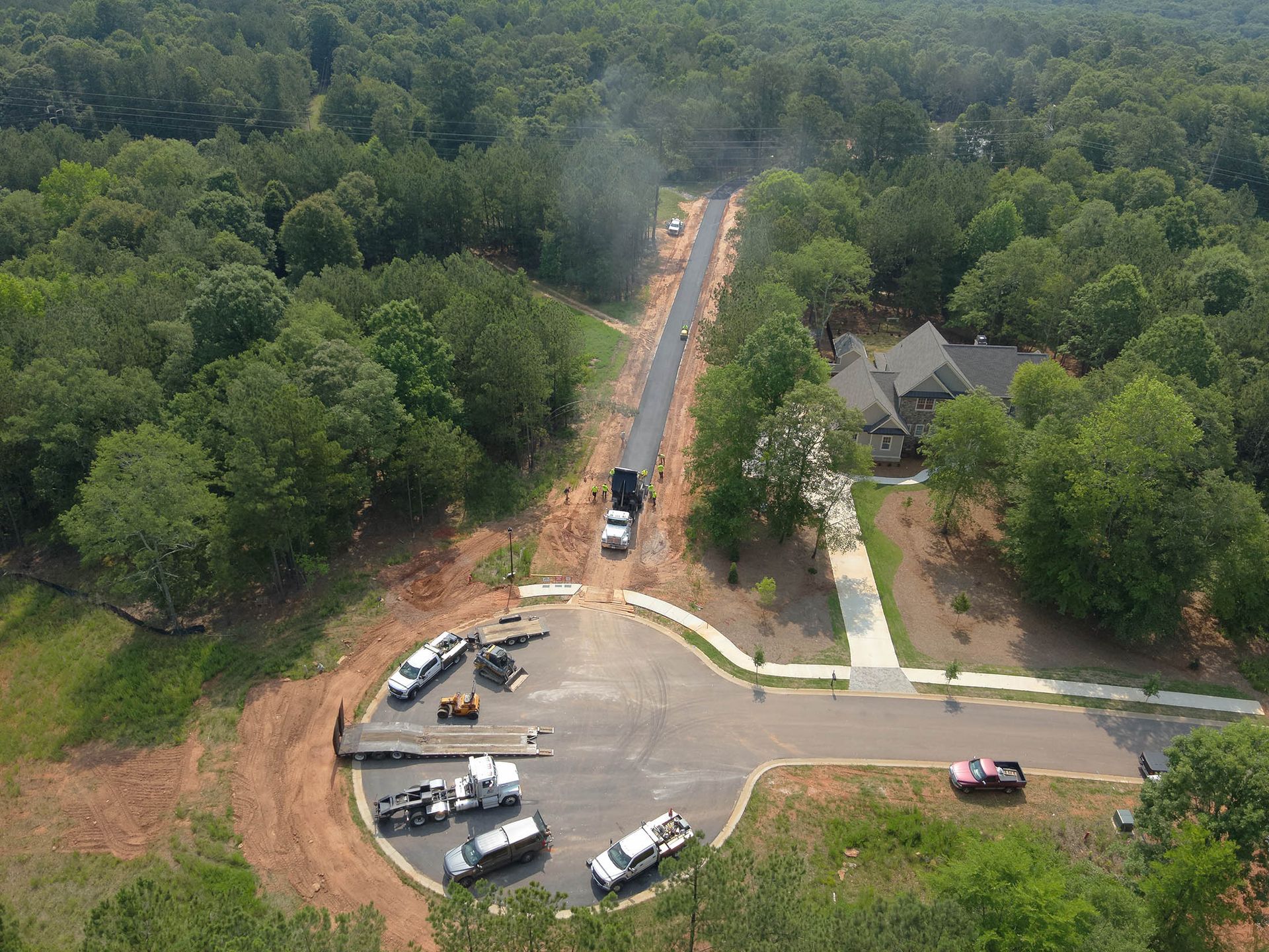 Aerial view of road paving in a wooded area, with construction vehicles and a house visible.