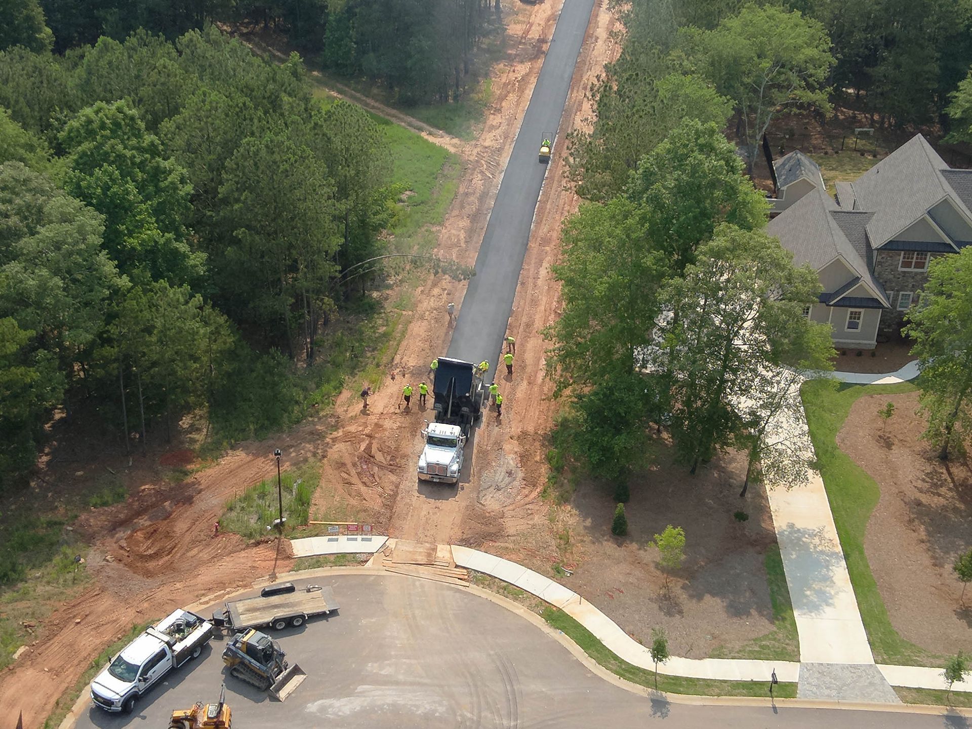 Road paving in progress, asphalt being laid by truck on a new road, surrounded by trees and a house.