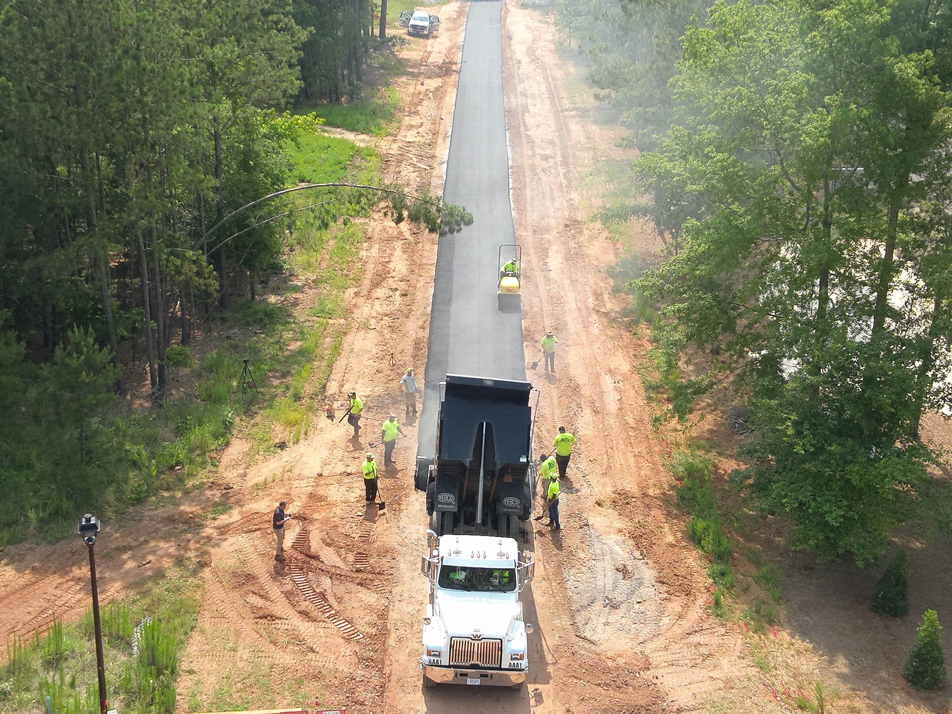 Dump truck unloading asphalt, workers in safety vests, surrounded by dirt and trees.
