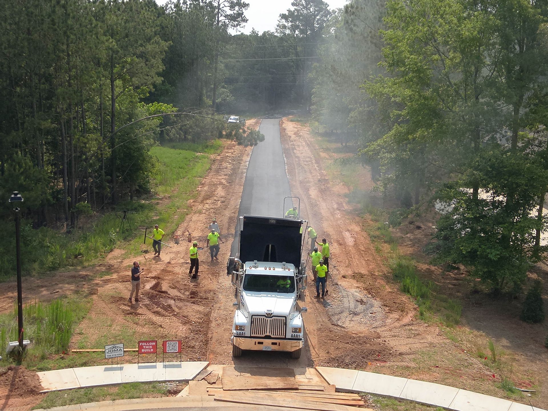 A dump truck unloading asphalt on a newly paved road. Workers in safety vests are present.
