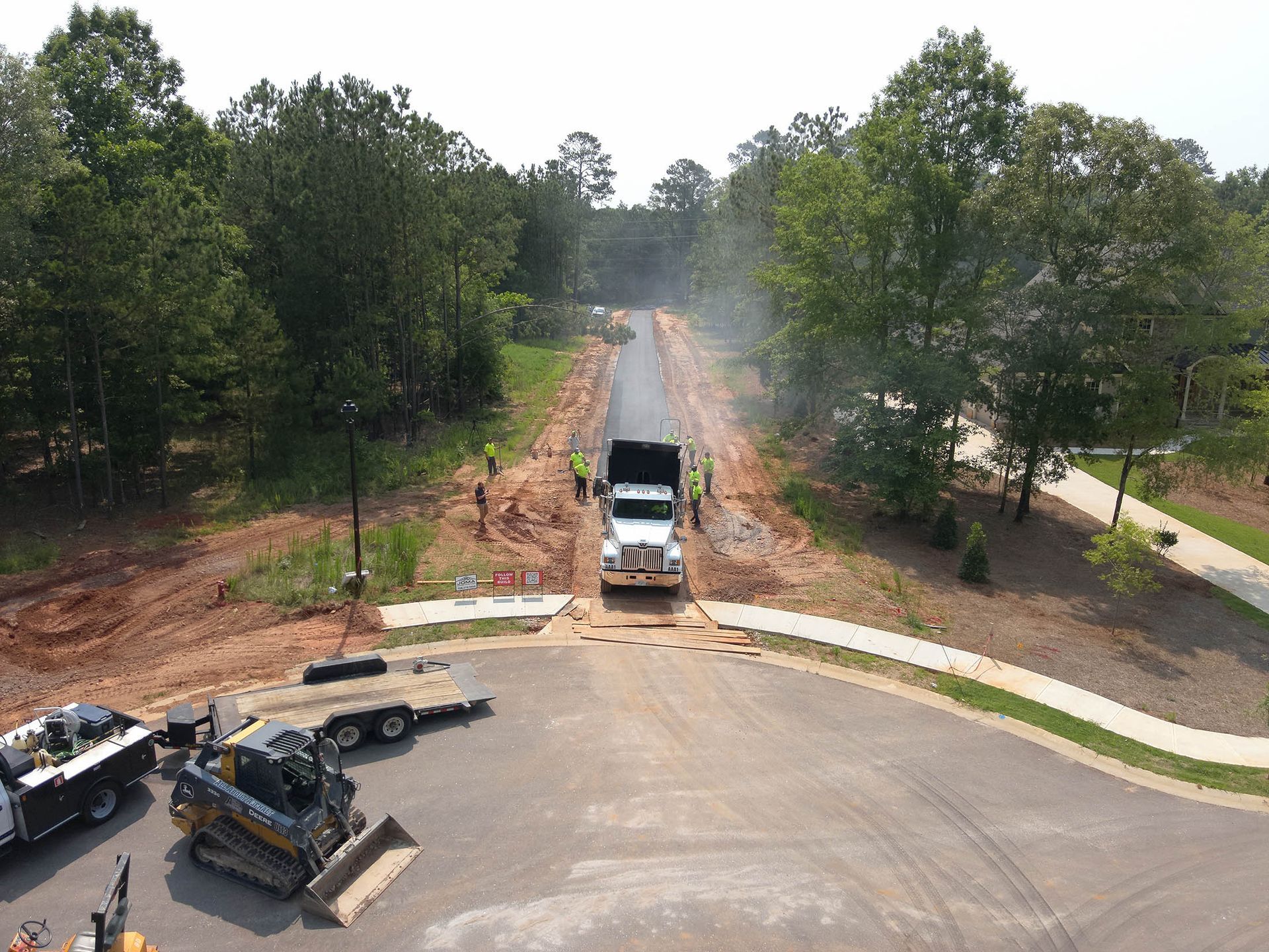 Truck unloading asphalt on a new road, with workers, equipment, and trees in the background.