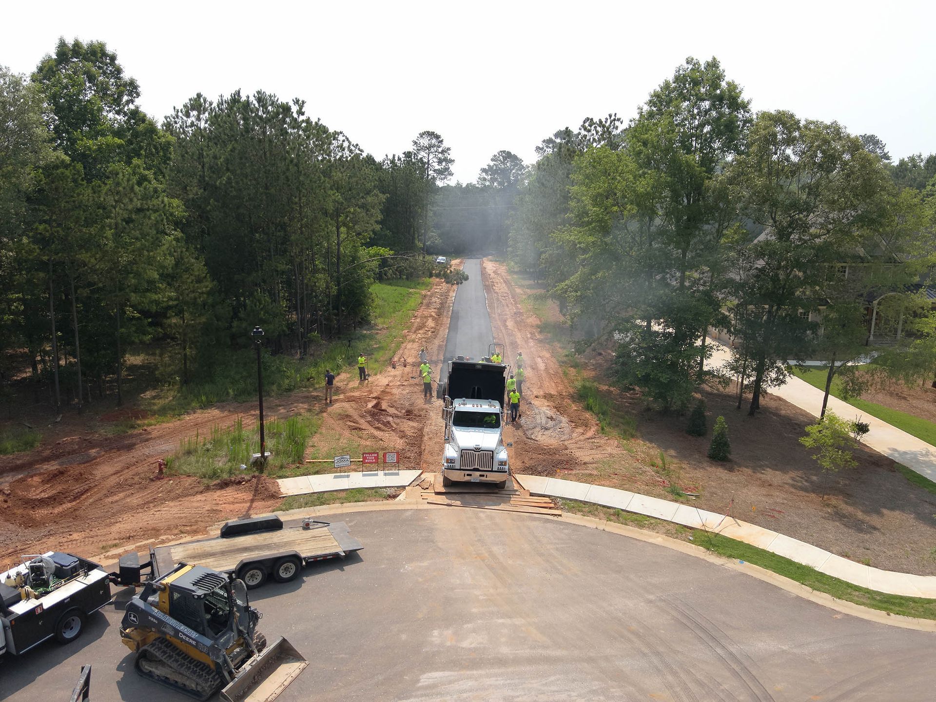 Truck unloading asphalt on a newly paved road, with workers, trees, and other construction equipment visible.