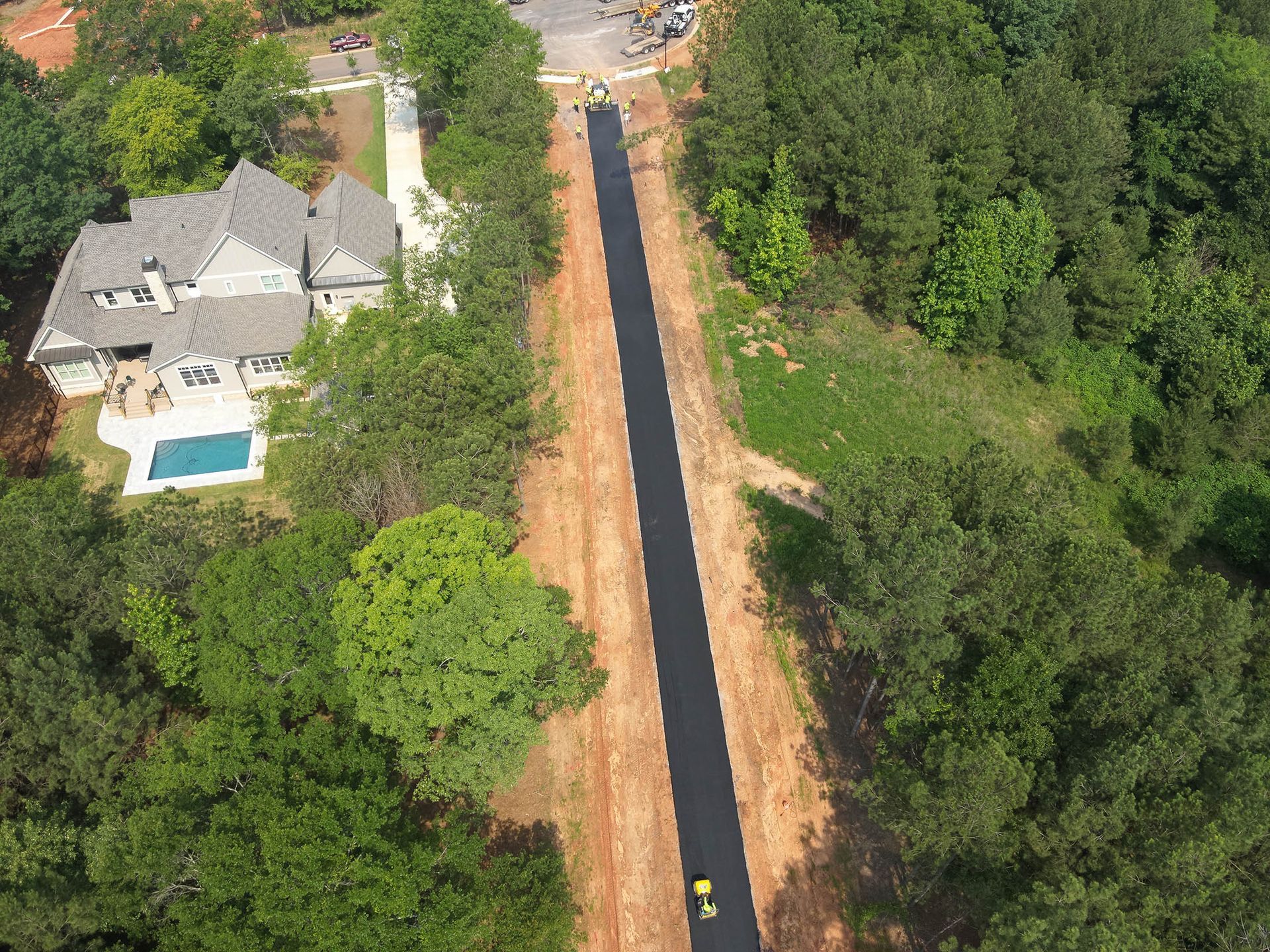 Aerial view of a paved pathway under construction through a wooded area, next to a house with a pool.