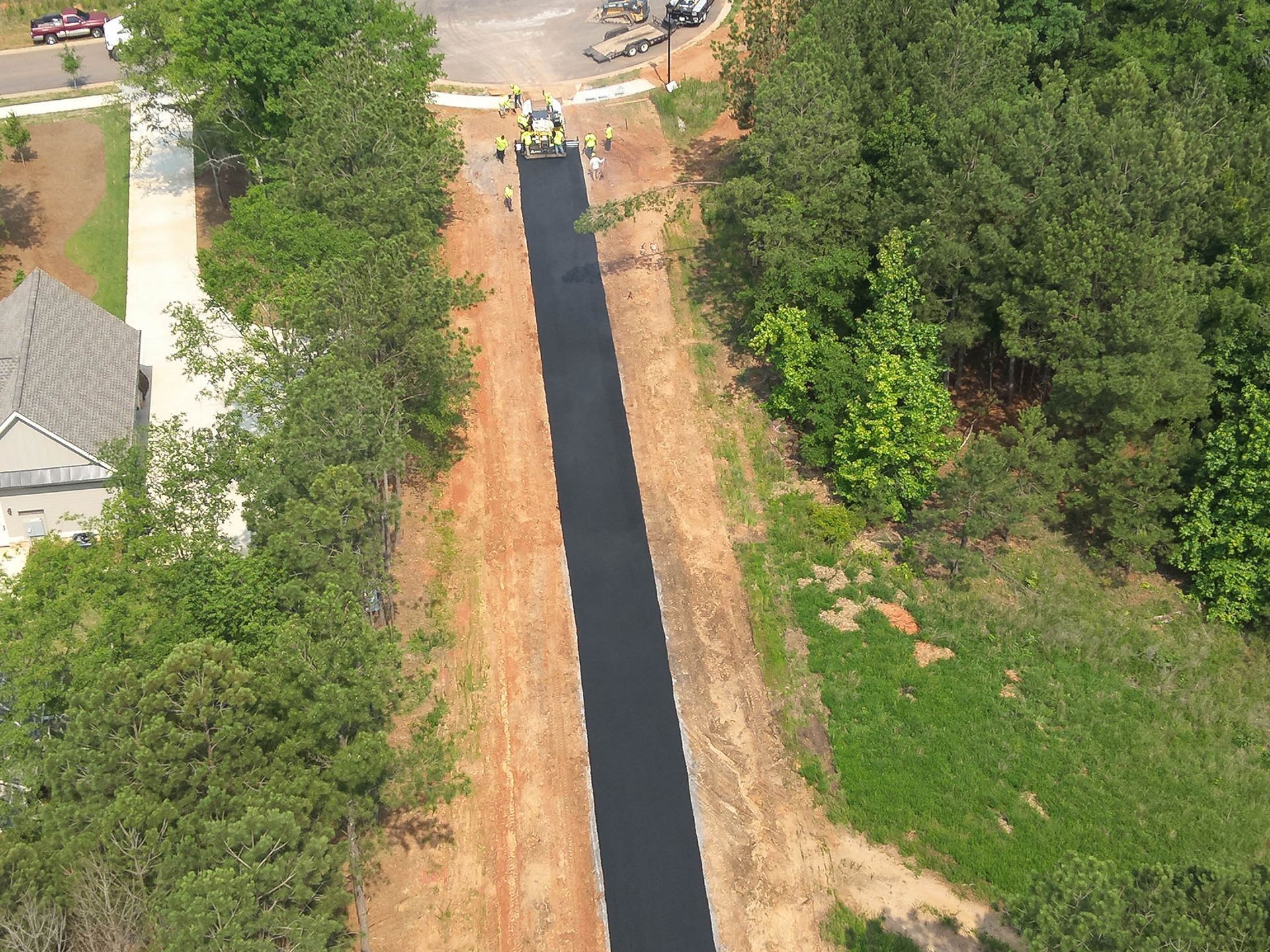 Newly paved road being constructed between trees, construction vehicles and workers visible.