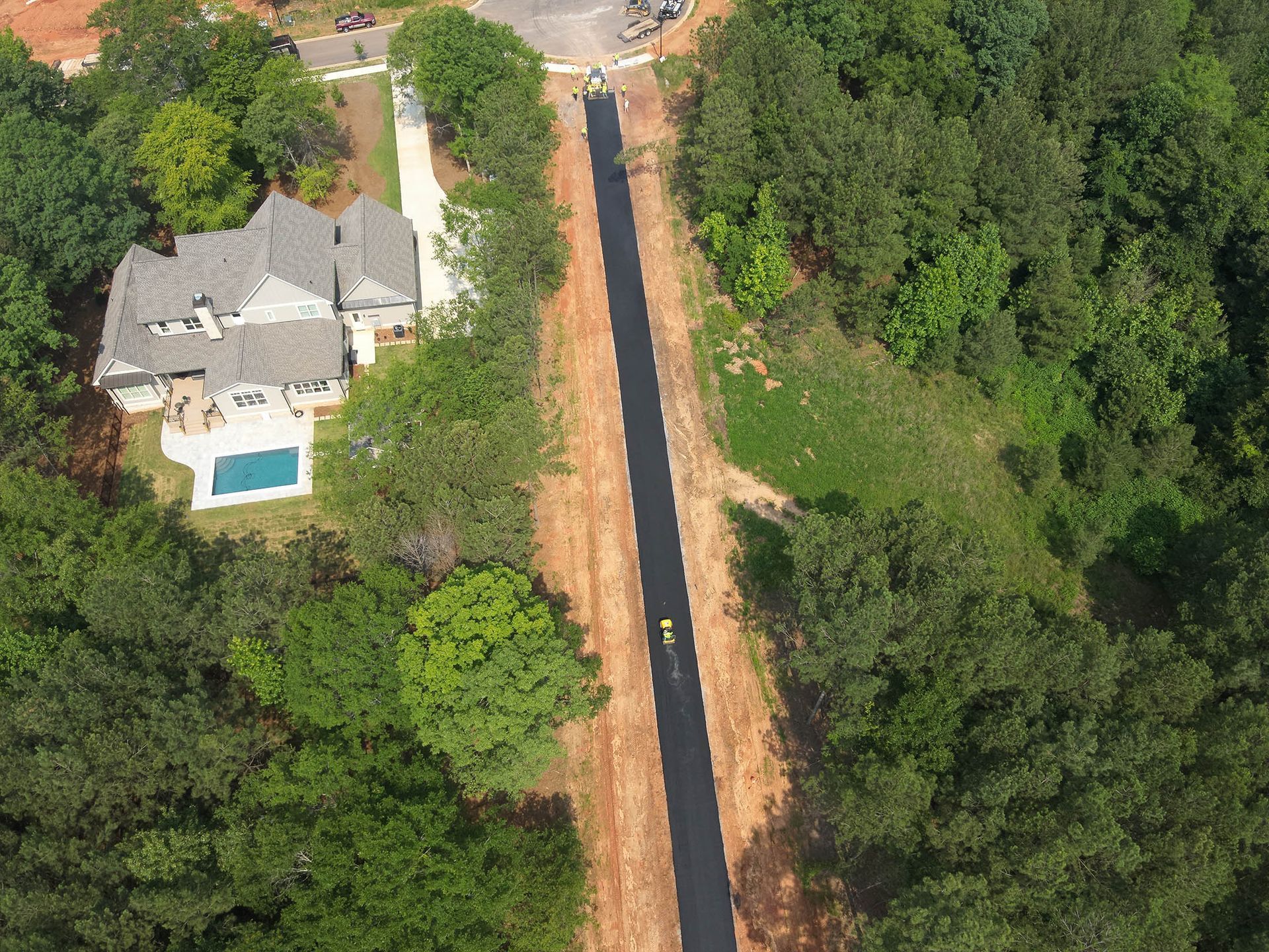 Aerial view of a long, newly paved road winding through trees and past a house with a pool.