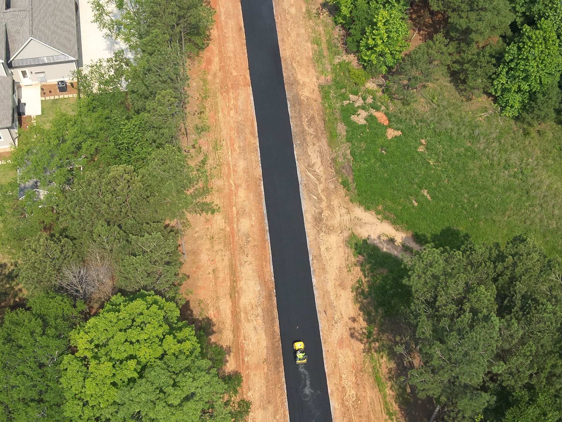 Aerial view of a paved path through a wooded area. A yellow vehicle drives down the path.