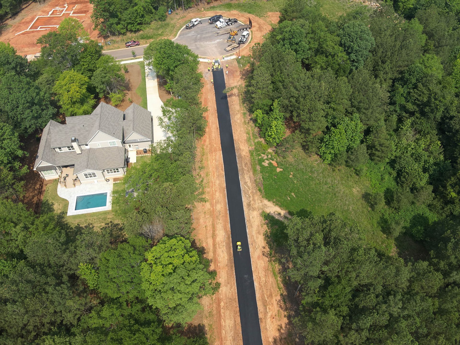 House with pool, long asphalt driveway, surrounded by trees and construction site.