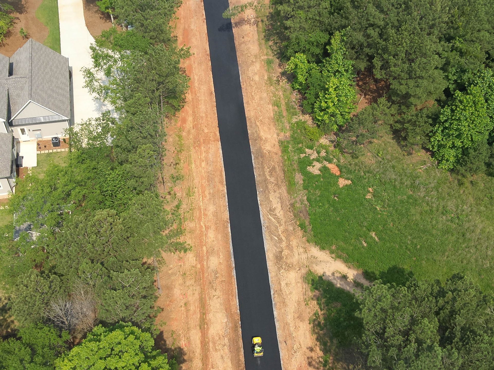 Aerial view of a paved pathway winding through a wooded area and past a house with a gray roof.