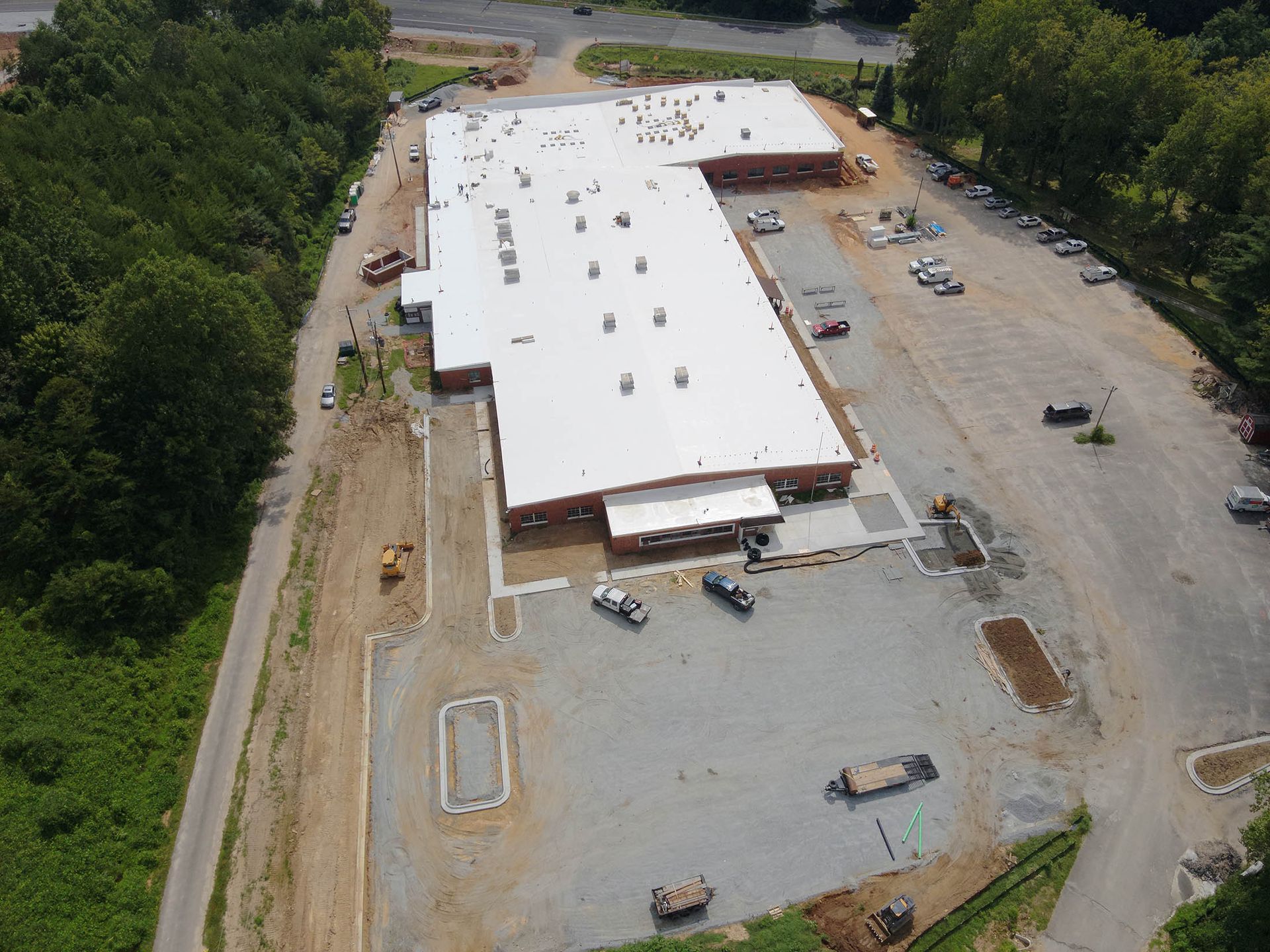 Aerial view of a large, white-roofed commercial building under construction, surrounded by trees and a partially paved parking area.