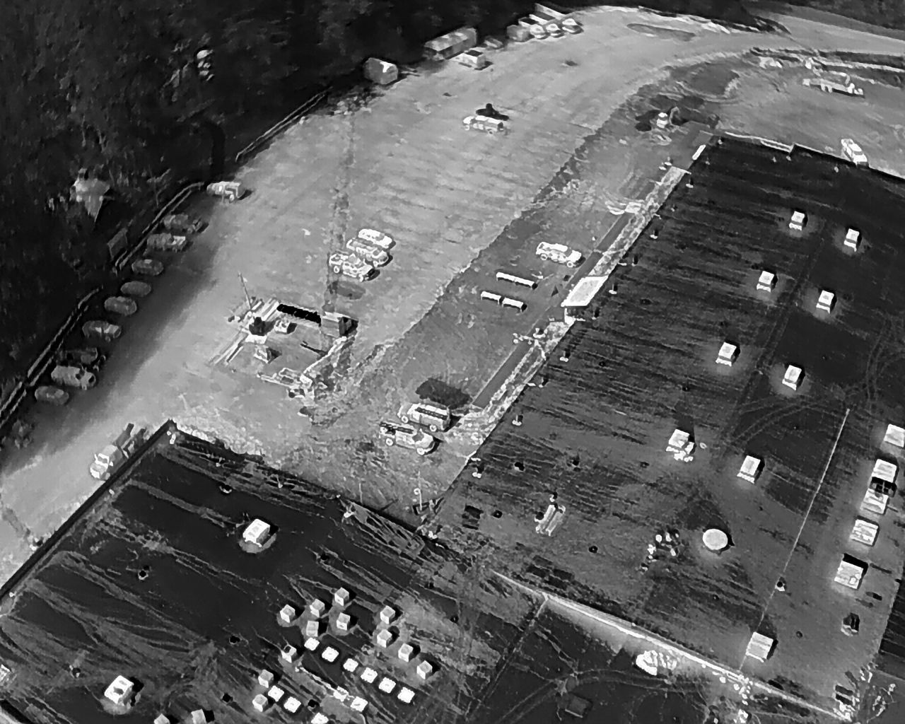 Overhead grayscale view of a warehouse area with buildings, parking, and forested areas.