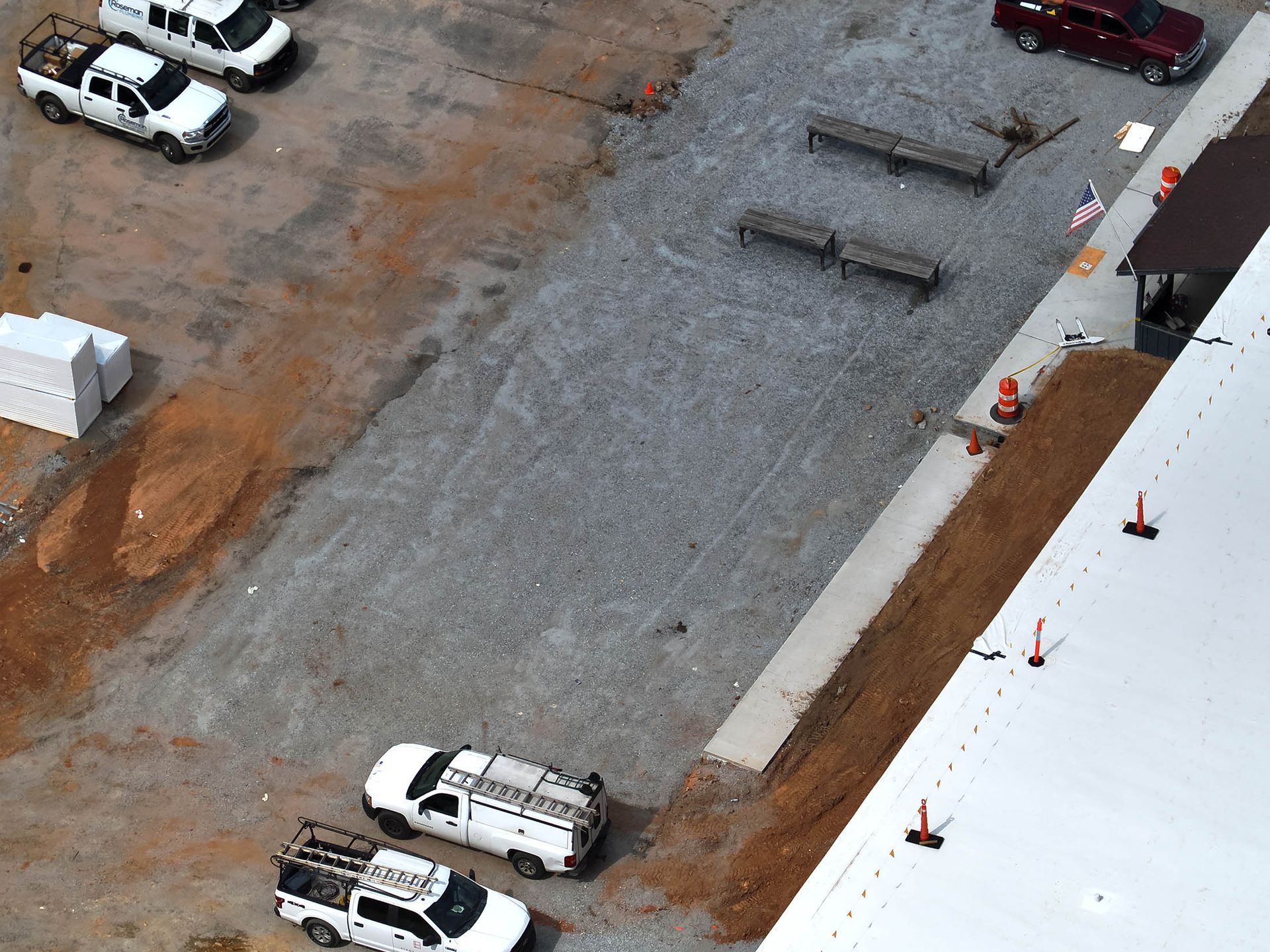 Aerial view of a construction site with parked trucks on gravel and dirt, benches, and a building with a white roof.