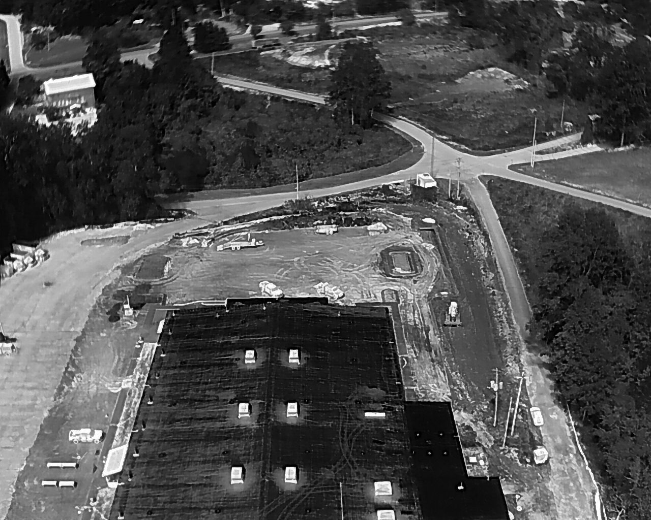 Aerial view of a flat, dark roof next to a construction site and intersection, surrounded by trees and grass.