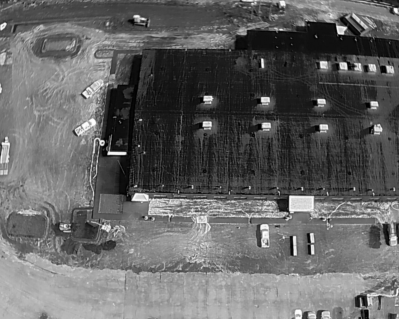 Overhead grayscale view of a flooded industrial building and surrounding area. Several vehicles are visible.