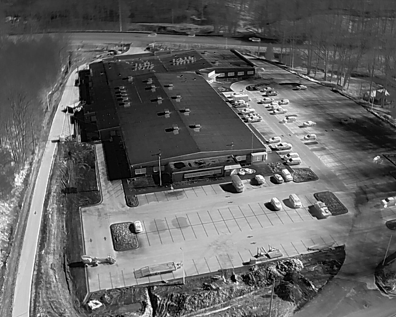 Aerial view of a commercial building with a large parking lot, surrounded by trees and a paved road.