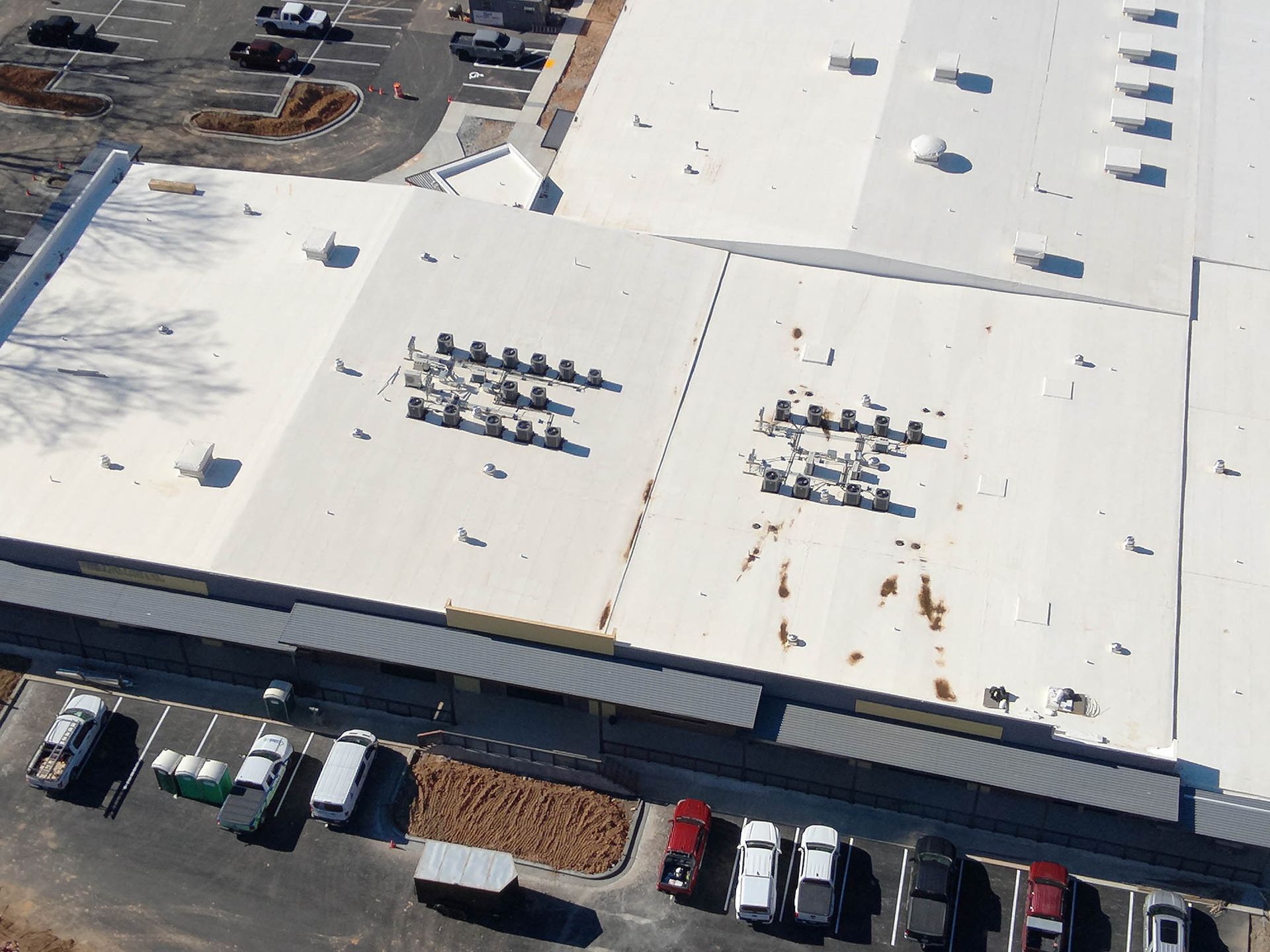 Overhead view of a large commercial building with a white roof, HVAC units, and a parking lot with vehicles.