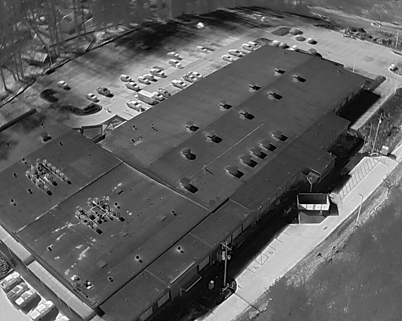 High-angle view of a large, dark building with a pocked roof and surrounding parking lot filled with cars.