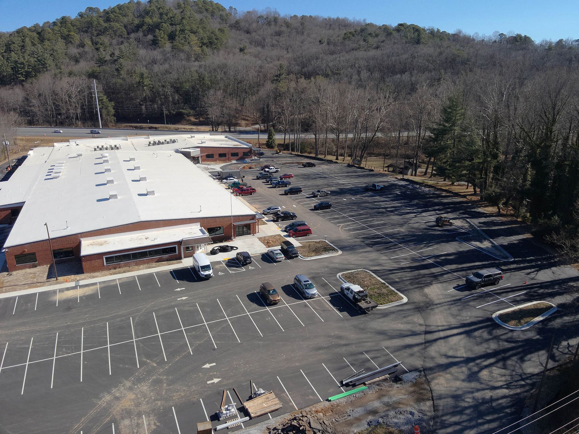 Aerial view of a commercial building with a large parking lot, cars, and surrounding trees on a sunny day.