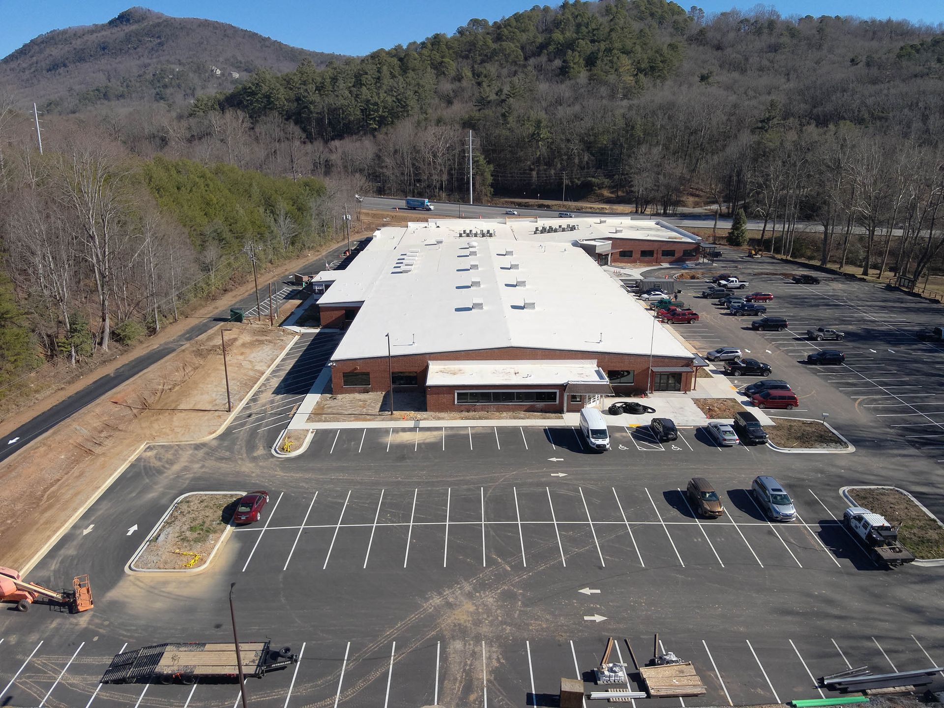 Aerial view of a long, brick-faced building with a large parking lot, mountains in the background.