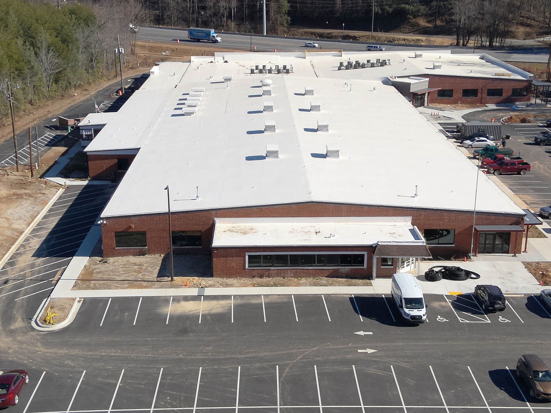 Aerial view of a long, brick building with a white roof, and a parking lot filled with cars.