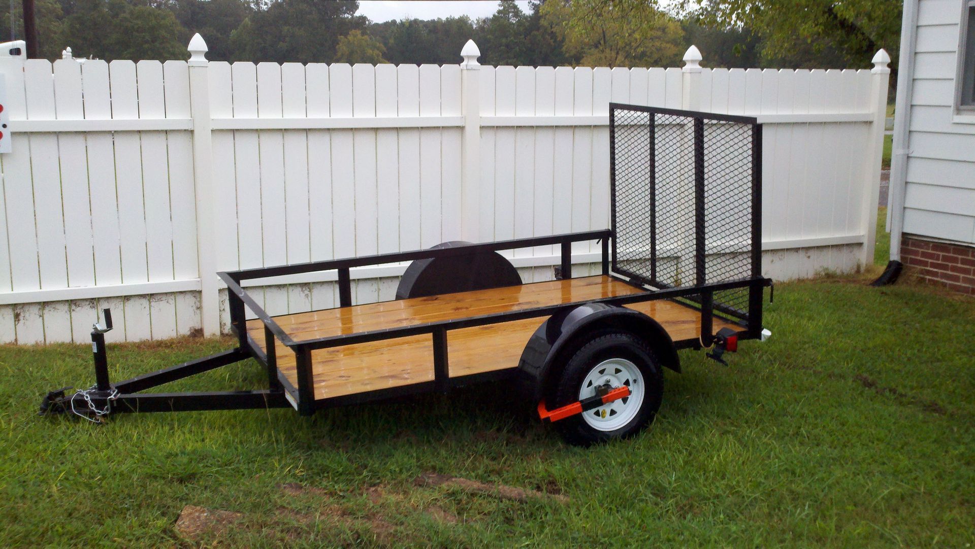 Black utility trailer on green grass in front of a white fence.