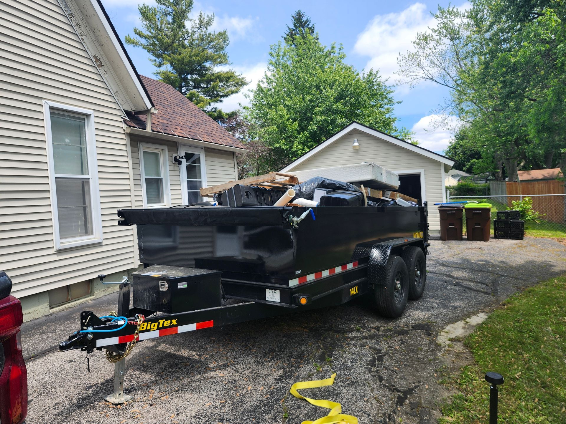 Black dumpster trailer parked in a driveway, next to a house and garage, loaded with debris.