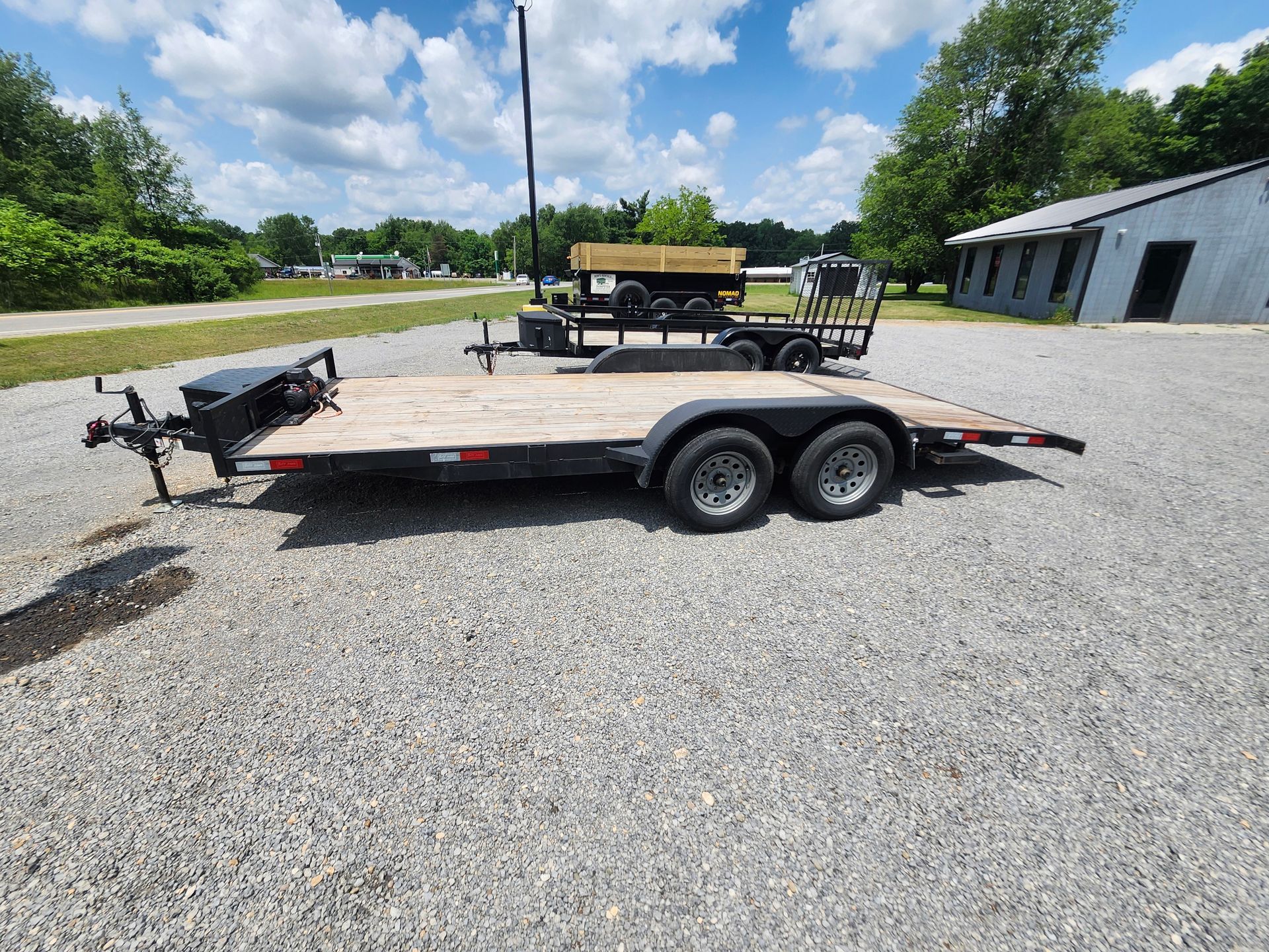 Two black trailers on gravel under a blue sky, one with a wooden deck, the other carrying equipment.
