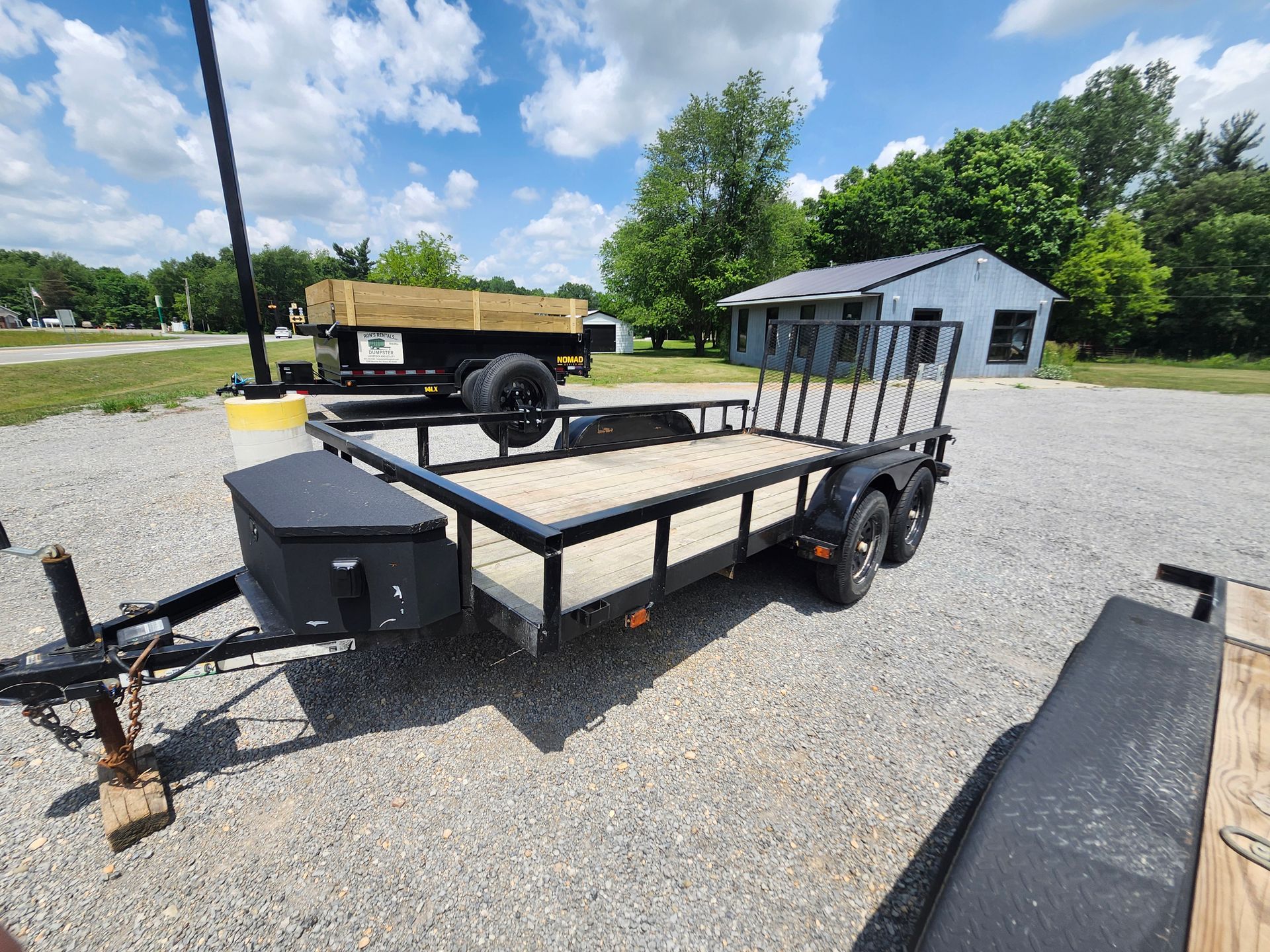 Black utility trailer with ramp, parked on gravel in front of a small building under a blue sky.