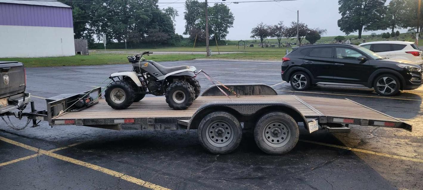 An ATV on a trailer is hitched to a black SUV parked on a paved lot. A purple building is in the background.