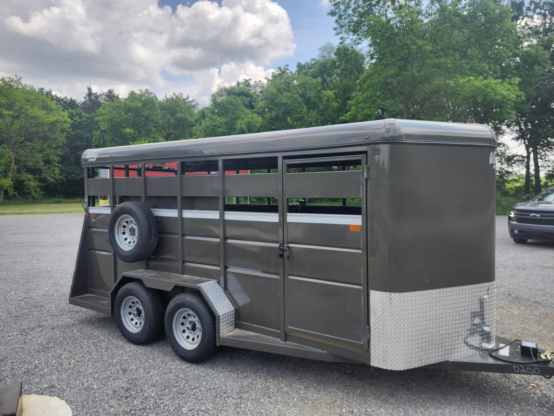 Dark grey stock trailer with two axles, spare tire, and open-slatted sides, parked on gravel.