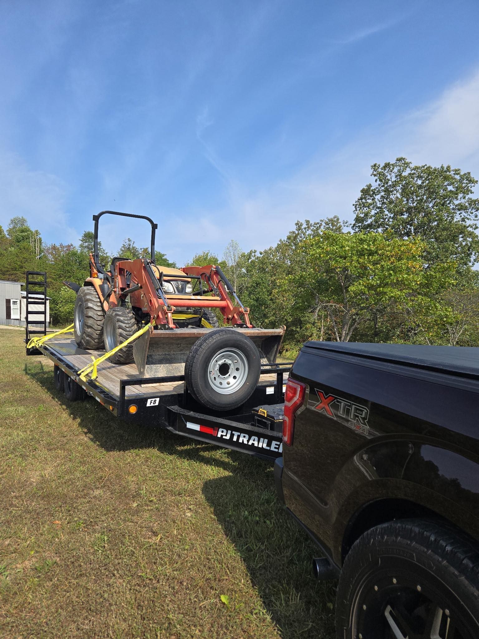 A black truck towing a trailer with an orange tractor on it, in a grassy field under a blue sky.