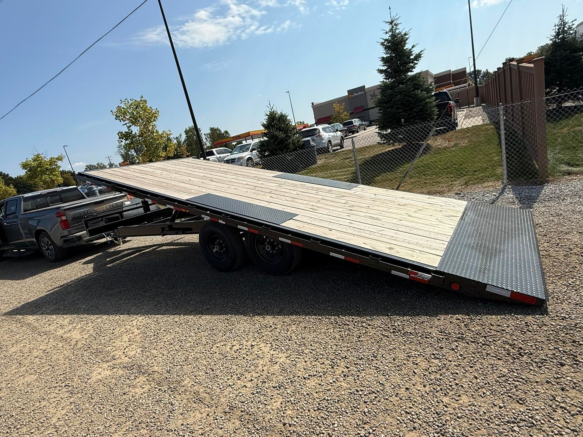 A gray pickup truck towing a large flatbed trailer on gravel, sunny day.