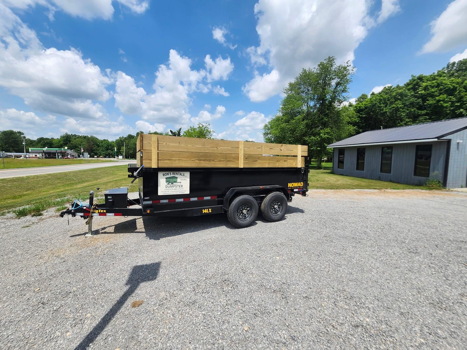 A black trailer with wooden sides parked on gravel. A sunny day with a building in the background.