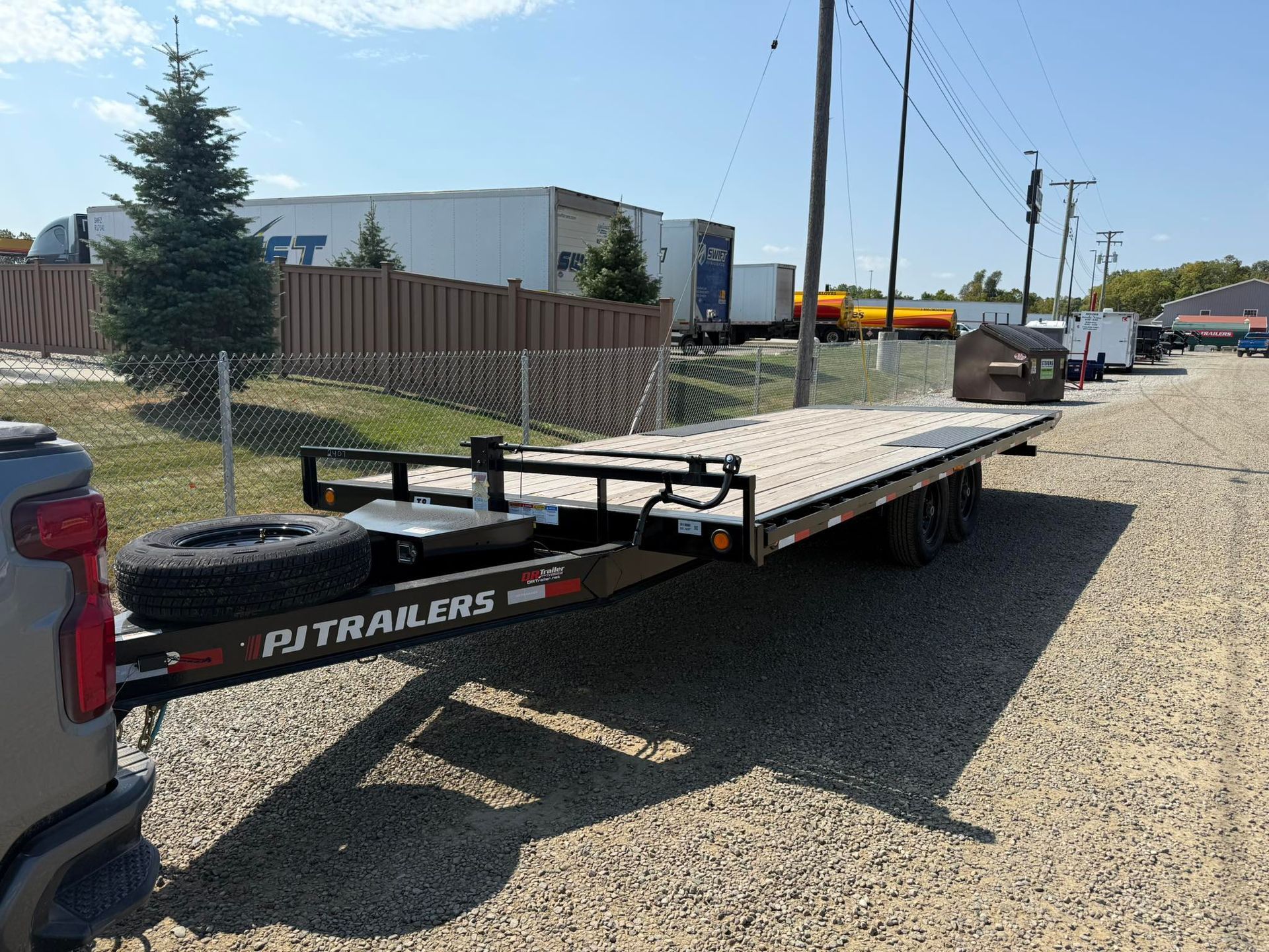 Black PJ Trailers flatbed trailer hooked to a truck on a gravel lot. Spare tire mounted on the trailer.