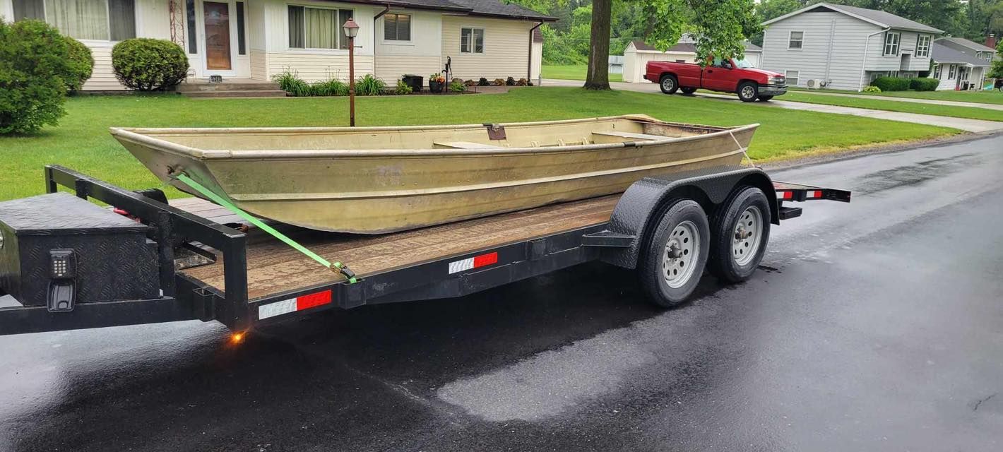 A boat on a trailer, parked on a wet street in a residential area. A red truck is in the background.