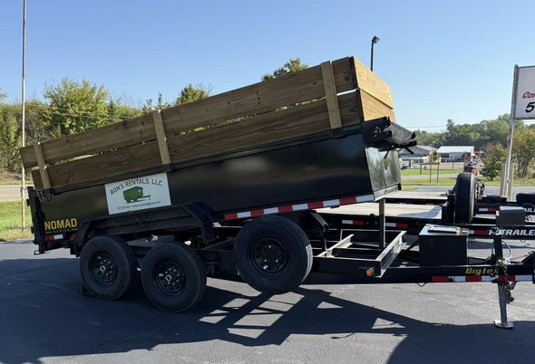 Black dump trailer with wooden sideboards raised on pavement, sunny day.