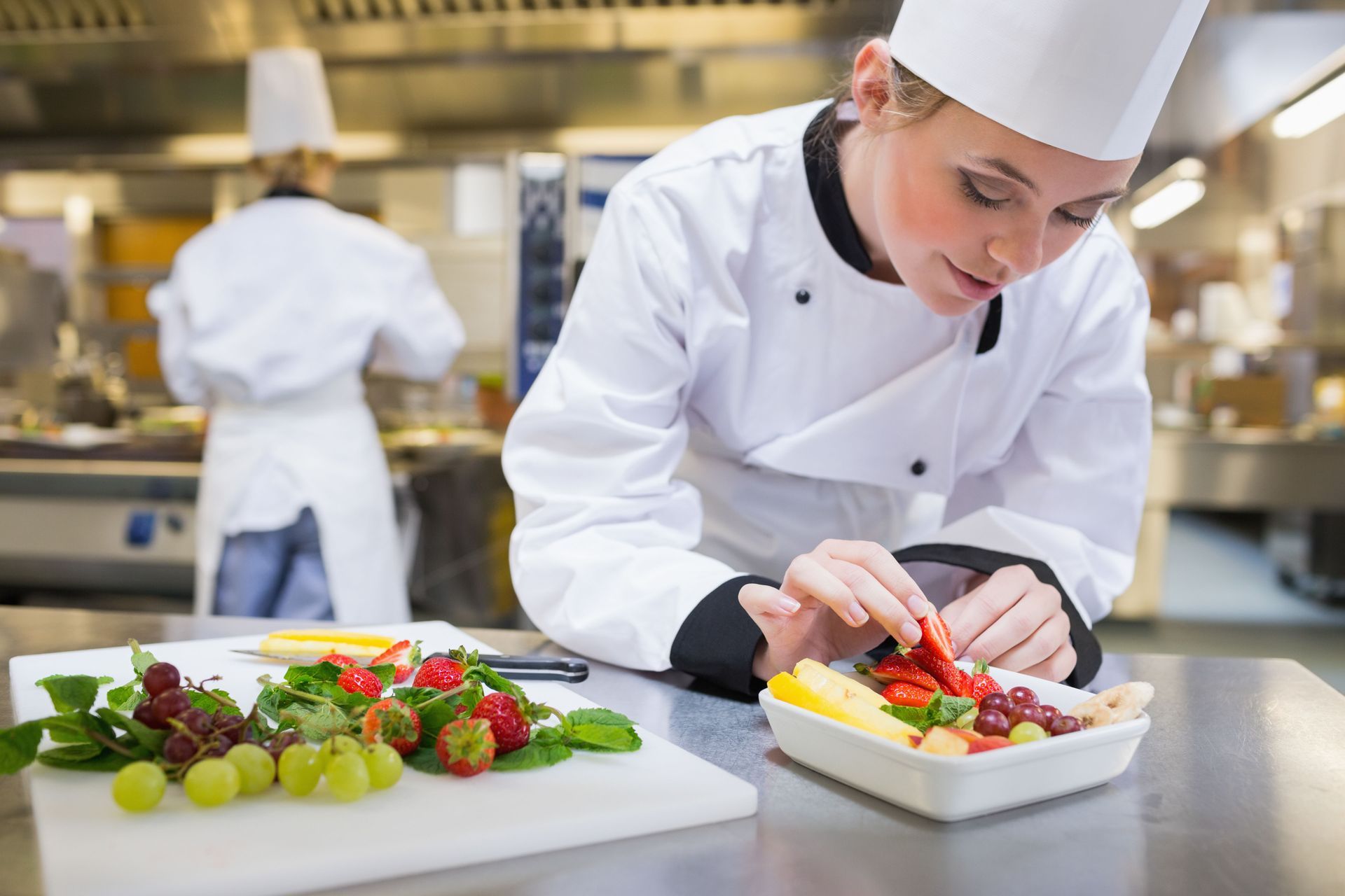 Chef garnishing fruit salad with grapes and strawberries in commercial kitchen.