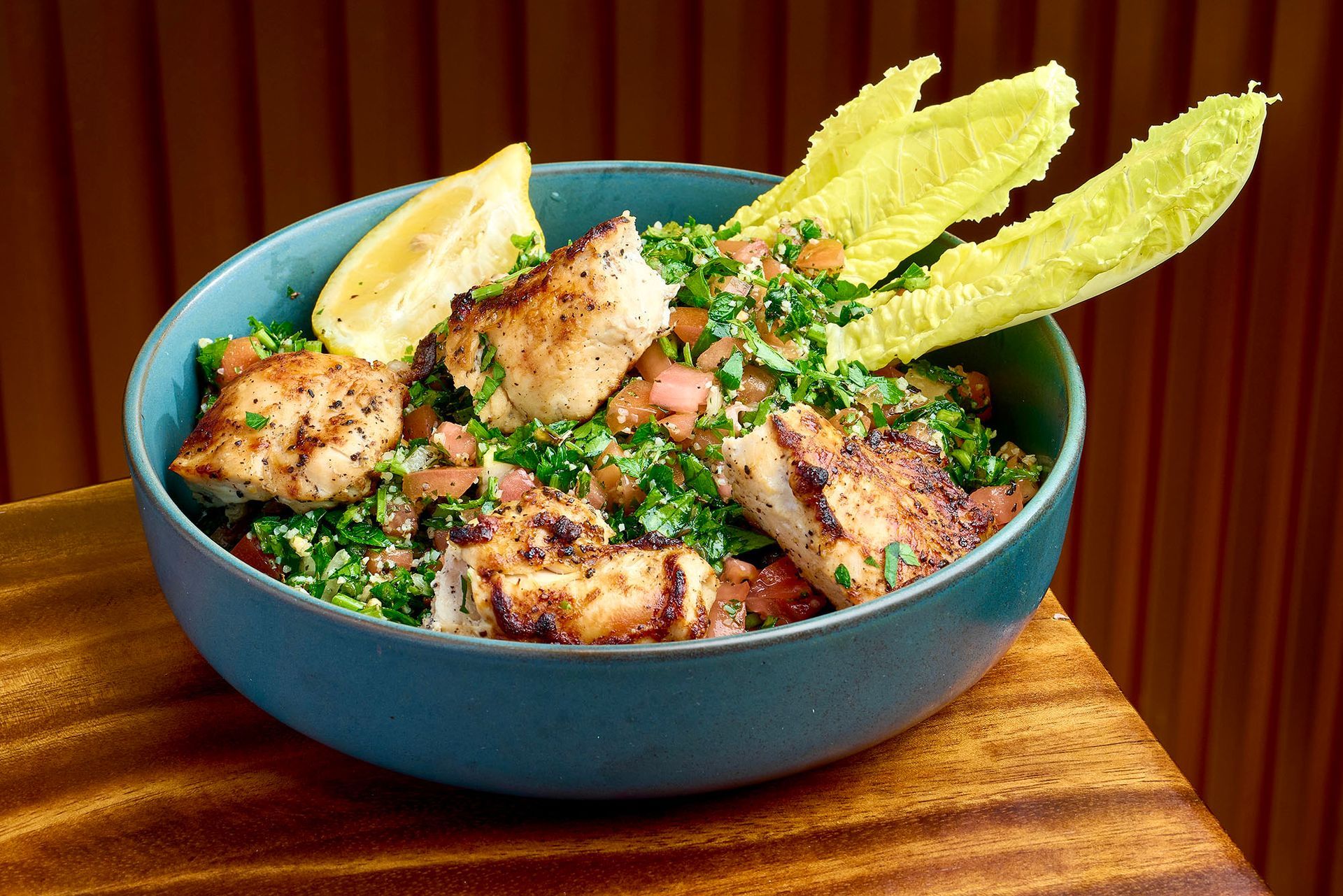Grilled chicken and tabbouleh salad in a blue bowl, with lemon and lettuce on a wooden surface.