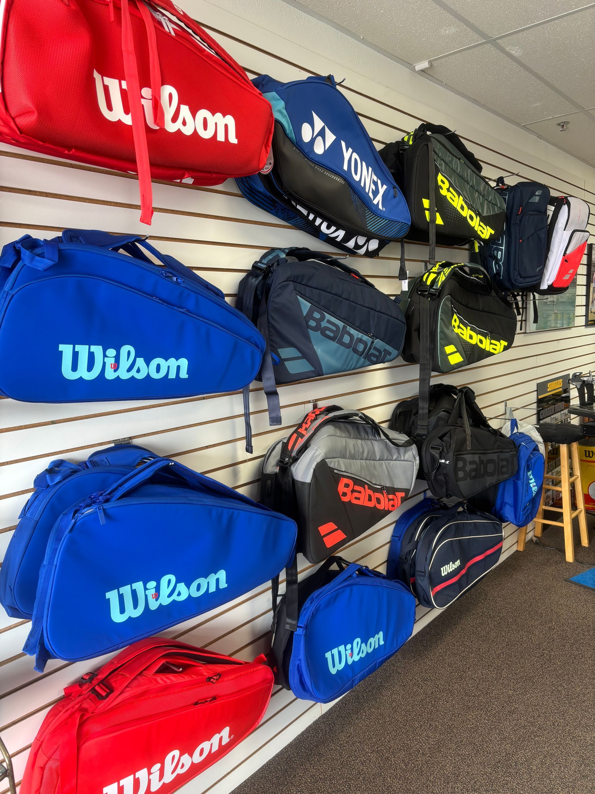 Multiple colorful tennis racquet bags by Wilson, Yonex, and Babolat hang on a slatted wall display in a sports shop.
