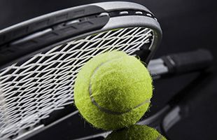 A yellow tennis ball rests against the strings of a tennis racket on a dark, reflective surface.