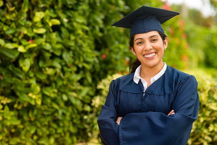 A woman in a graduation cap and gown is standing with her arms crossed and smiling.