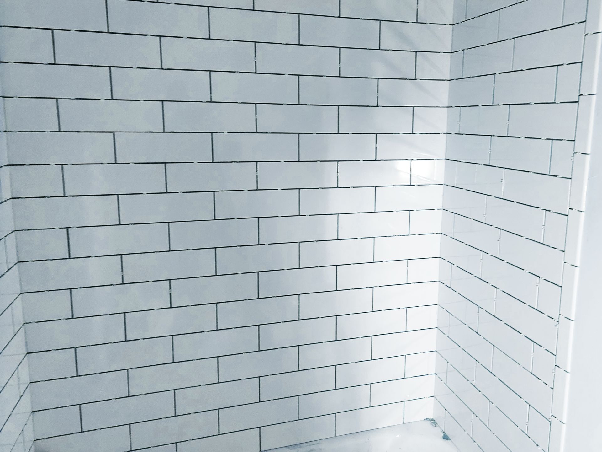 A close-up view of a shower wall featuring white, rectangular subway tiles with thin, dark grout lines.