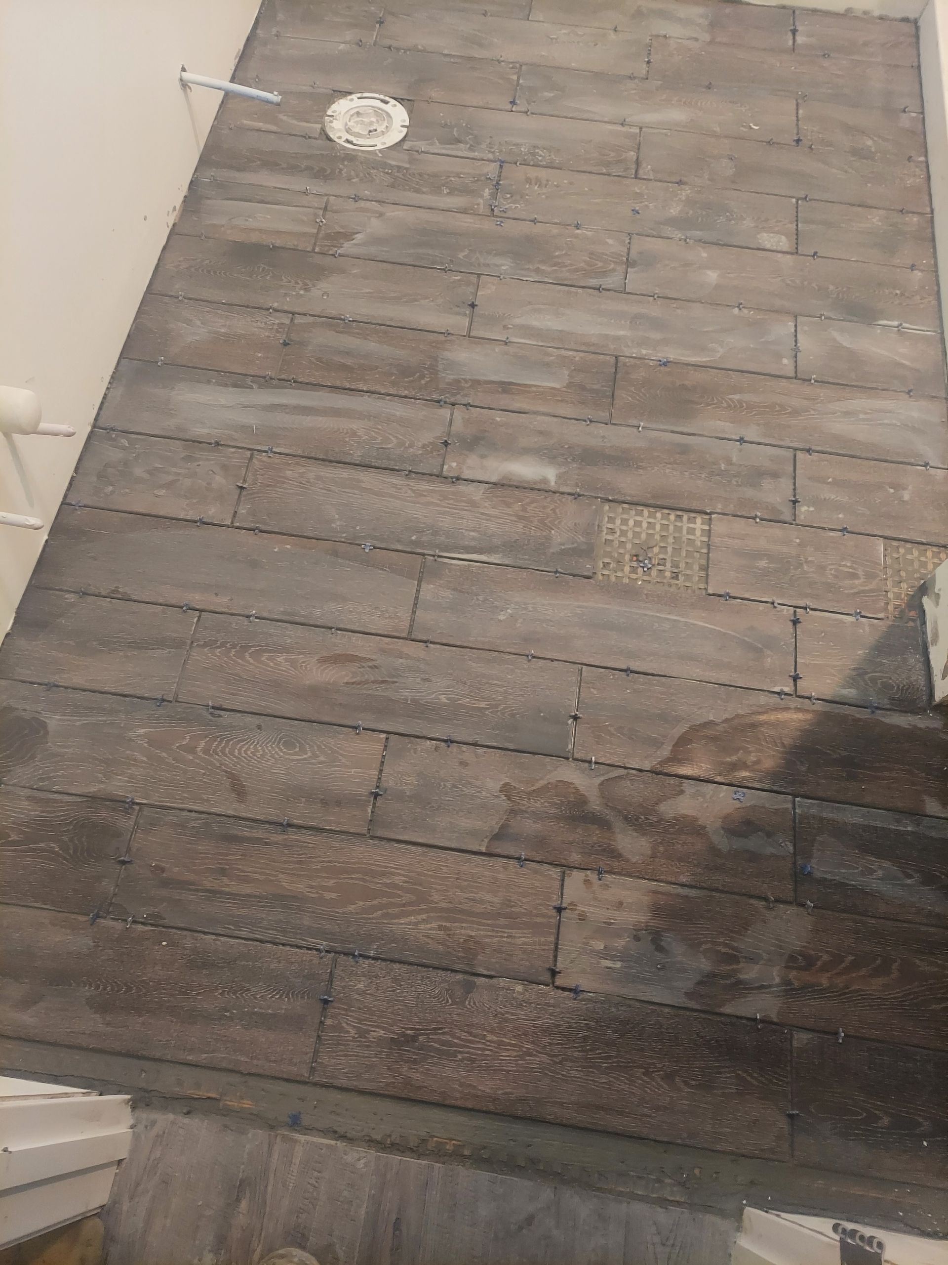 Gray rectangular floor tiles are installed in a running bond pattern in a room with a visible toilet flange and pipes.