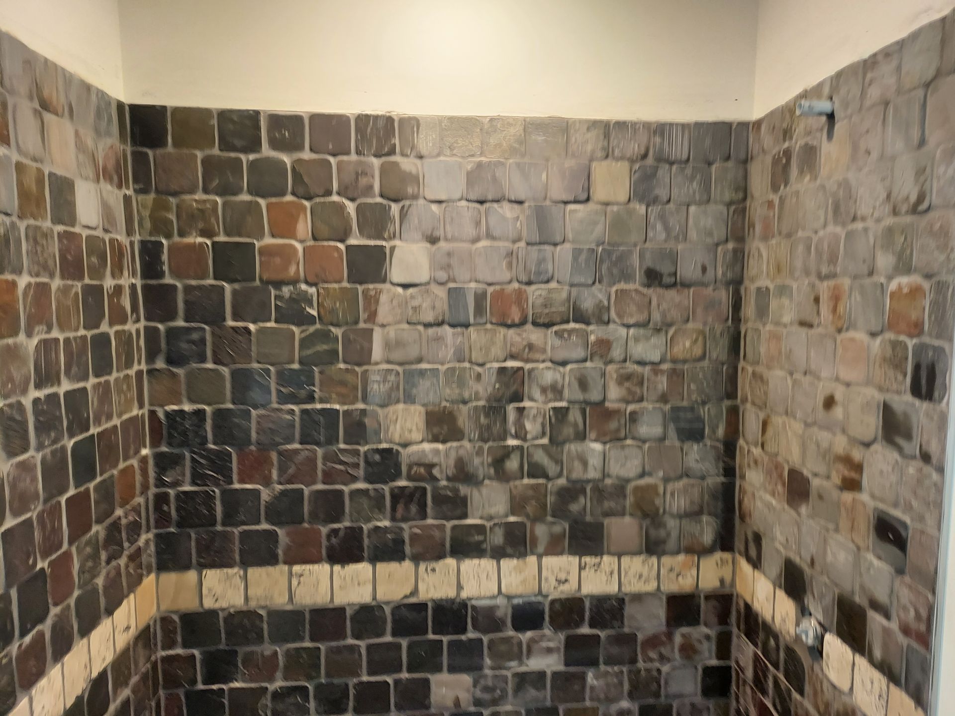 A shower wall tiled with multi-colored, square natural stones featuring a horizontal decorative row of light cream tiles.