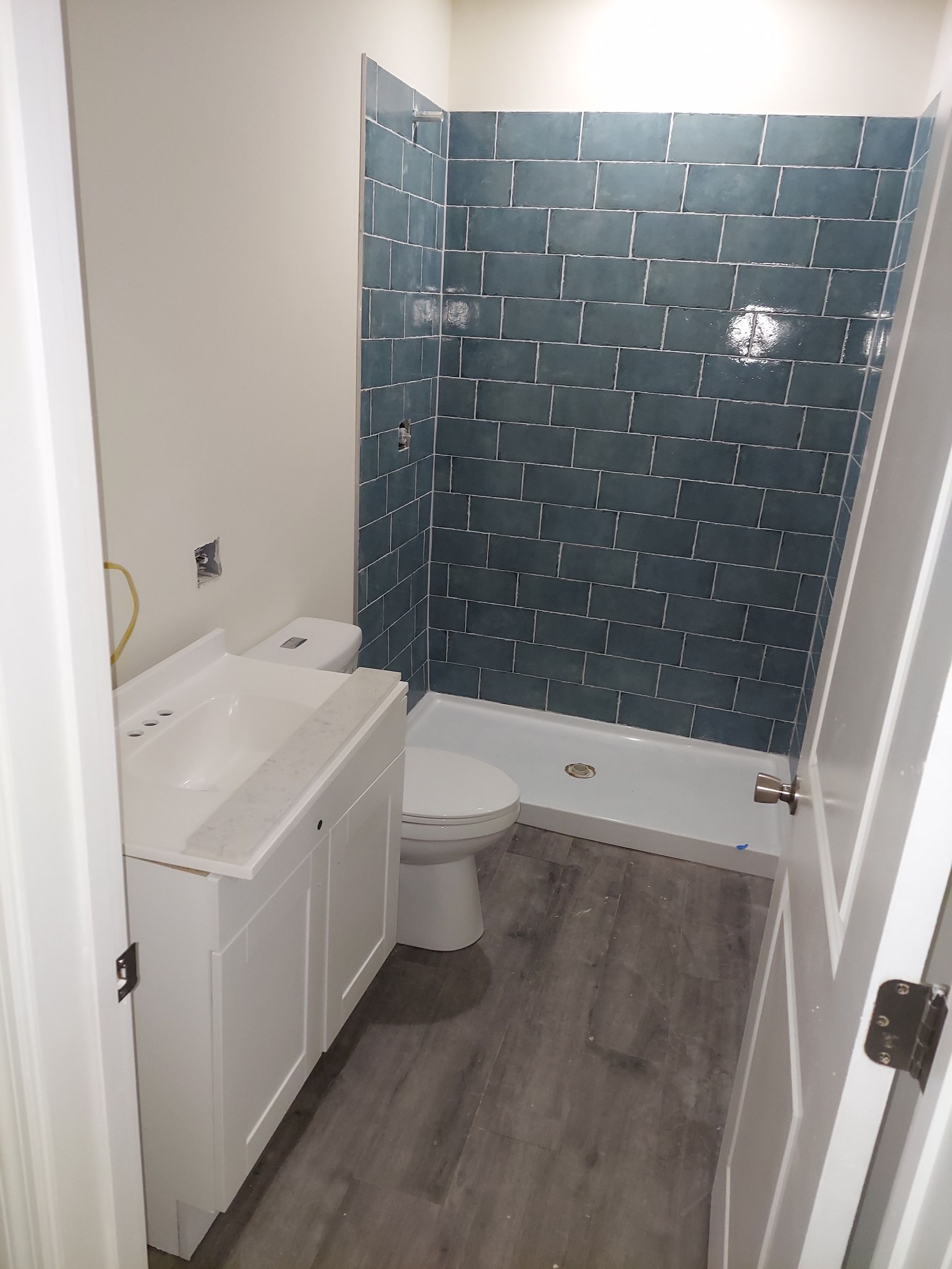 A small bathroom with a white vanity and toilet, gray wood-look flooring, and a dark blue tiled shower surround.