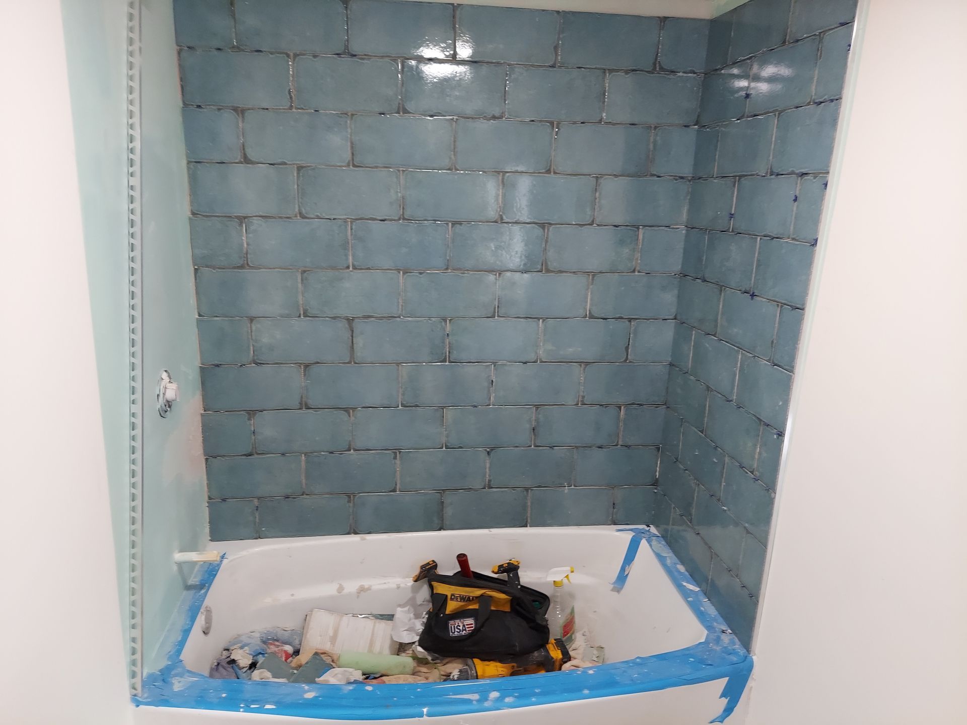 A bathtub in an alcove with blue, brick-patterned wall tiles and protective blue painter's tape along the edges.