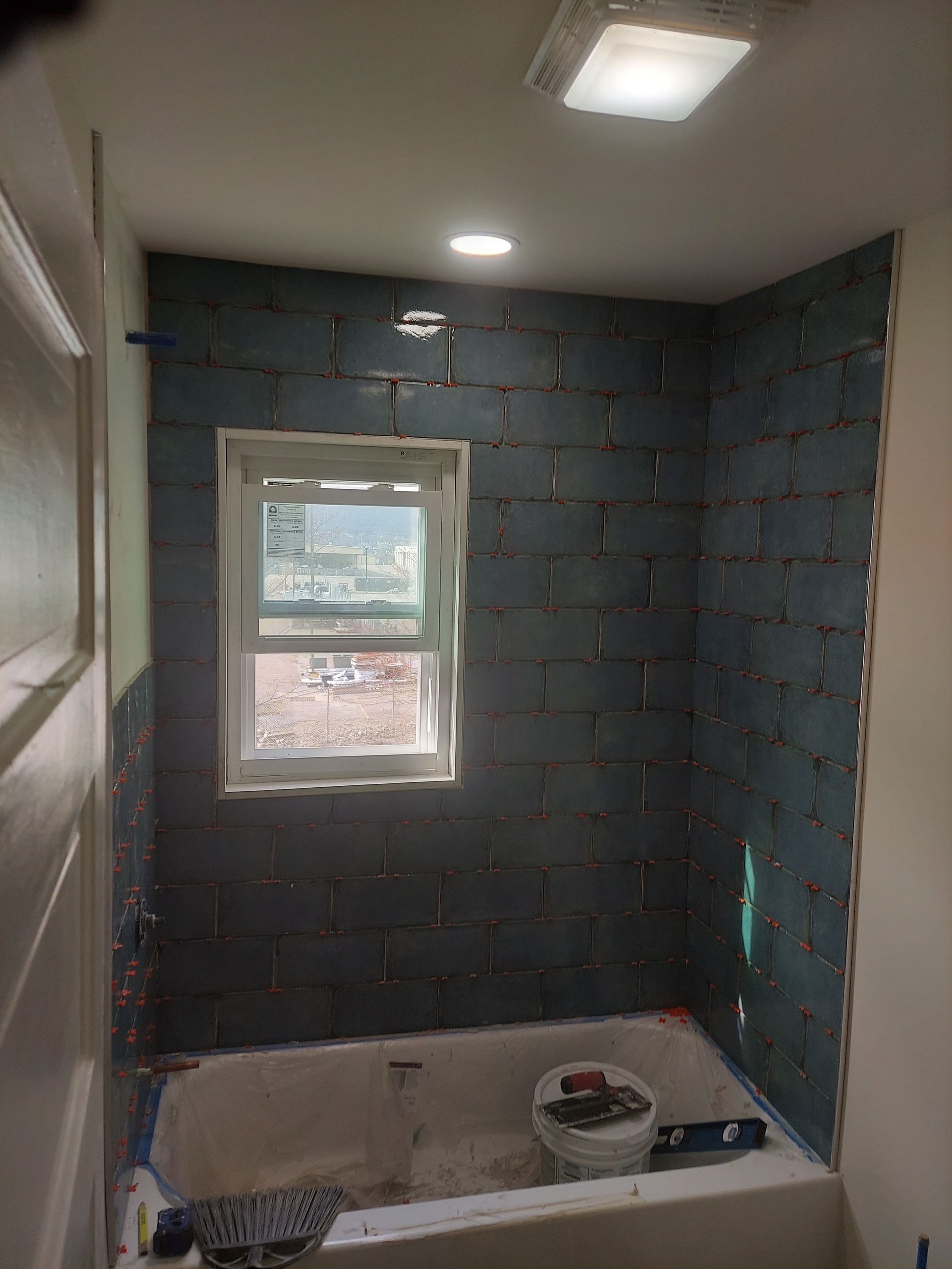 A bathroom shower stall under renovation, featuring grey cement board walls surrounding a window and a white bathtub.